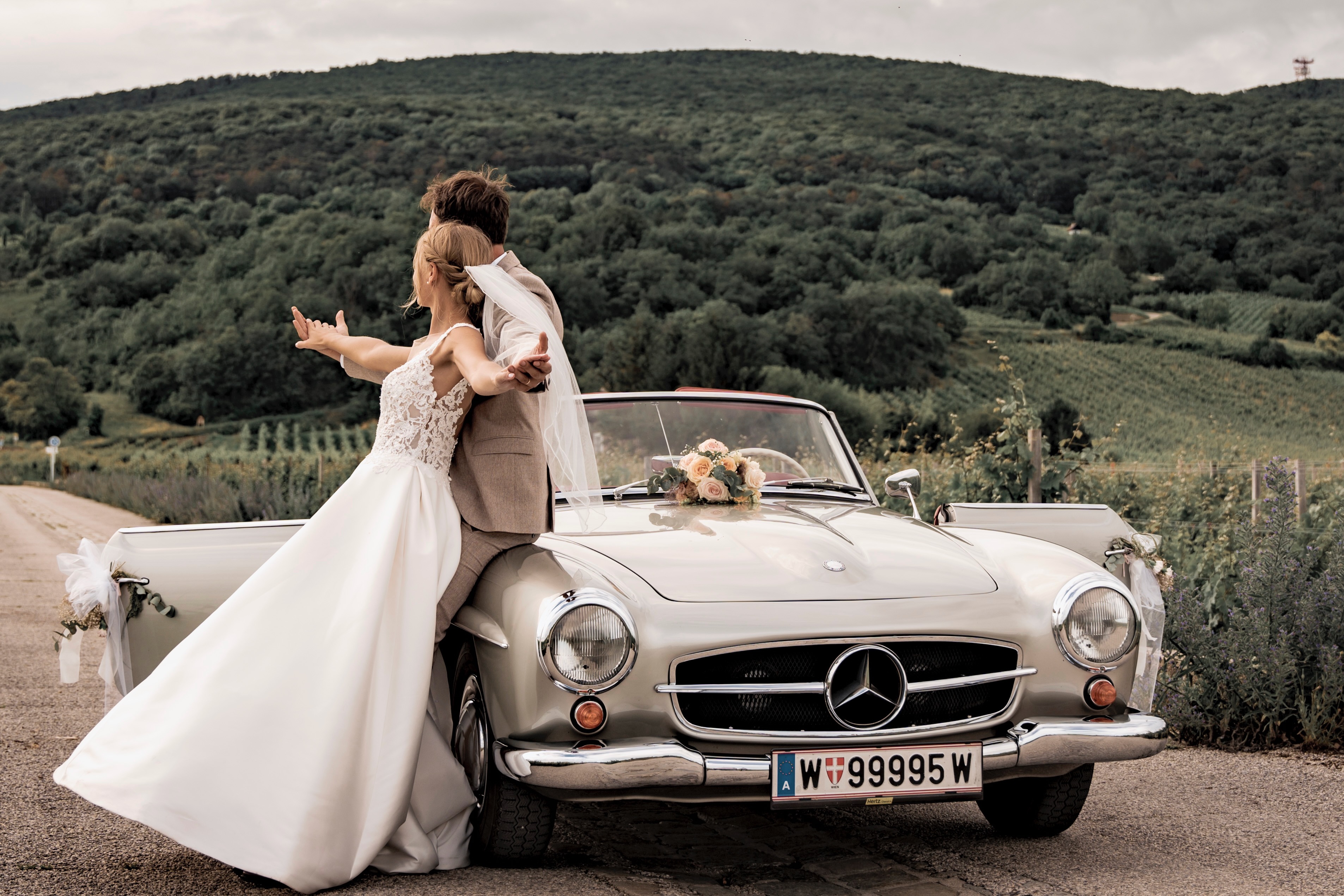 Brautpaar am Hochzeitstag an einem Oldtimer-Cabrio vor weiter Hügellandschaft