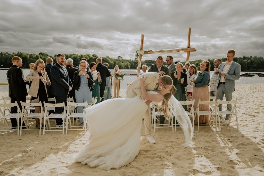 Brautpaar küsst sich am Strand — Hochzeitsmoment mit Gästen und Seifenblasen
